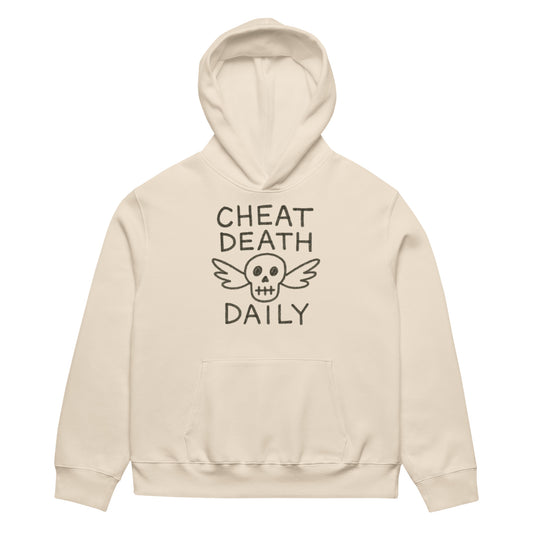 Person wearing a beige hoodie with Cheat Death Daily' text and skull  design. By Highway Division HWY DIV