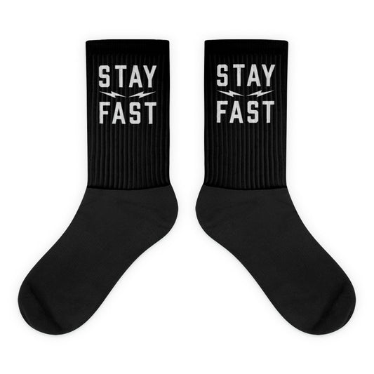 A pair of black crew-length socks from Highway Division is displayed on a clean, neutral background or worn by a model styled in rugged, streetwear-inspired clothing. The socks feature bold white woven text across the top cuff that reads “STAY FAST” in all capital letters. The lettering is clear, sharp, and intentionally placed to remain visible just above the ankle when worn with sneakers or boots, making a bold statement with every step. The socks are made from a high-quality cotton blend