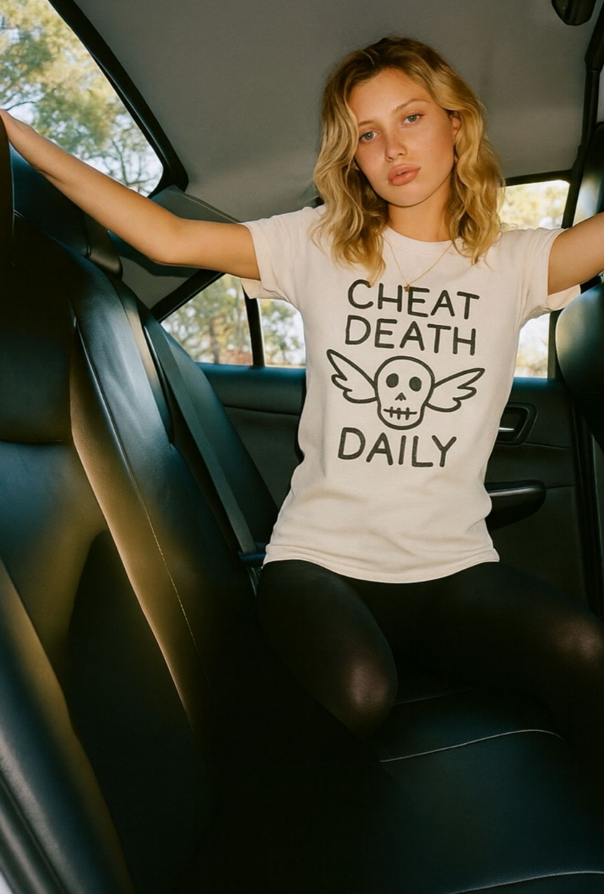 A blonde woman with loose, wavy hair is sitting in the back seat of a car, wearing a black t-shirt that reads “CHEAT DEATH DAILY” in bold, white capital letters across the chest. The shirt is part of the Highway Division collection and has a minimalist, rebellious aesthetic. The woman is facing slightly toward the window, with natural light highlighting her hair and the texture of the shirt. She has a confident, calm expression, exuding a sense of edge and defiance. The interior of the car is slightly 

