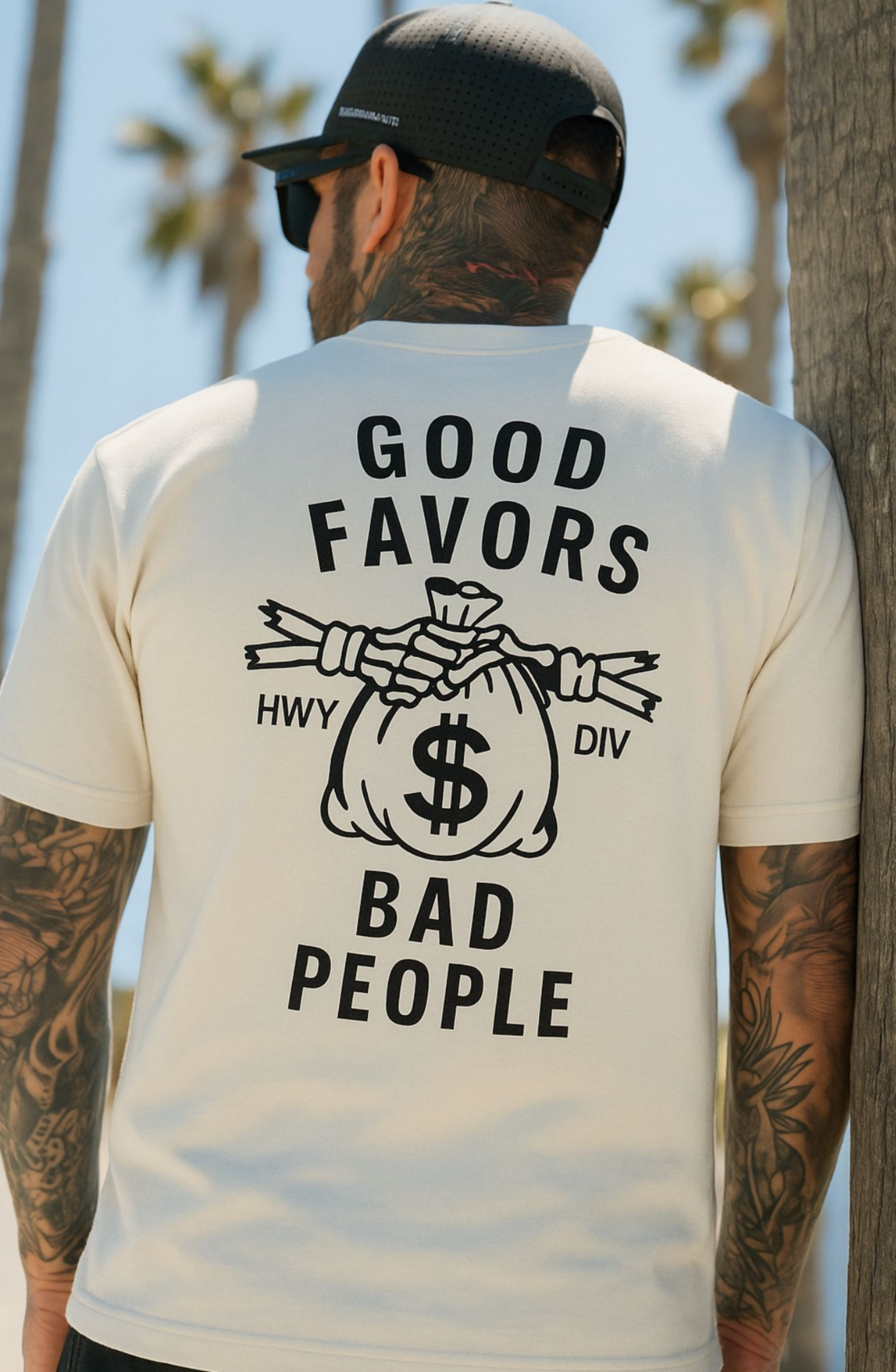 Model wearing vintage white Highway Division t-shirt with bold "GOOD FAVORS BAD PEOPLE" front print; rebellious streetwear with a gritty, anti-mainstream aesthetic.
