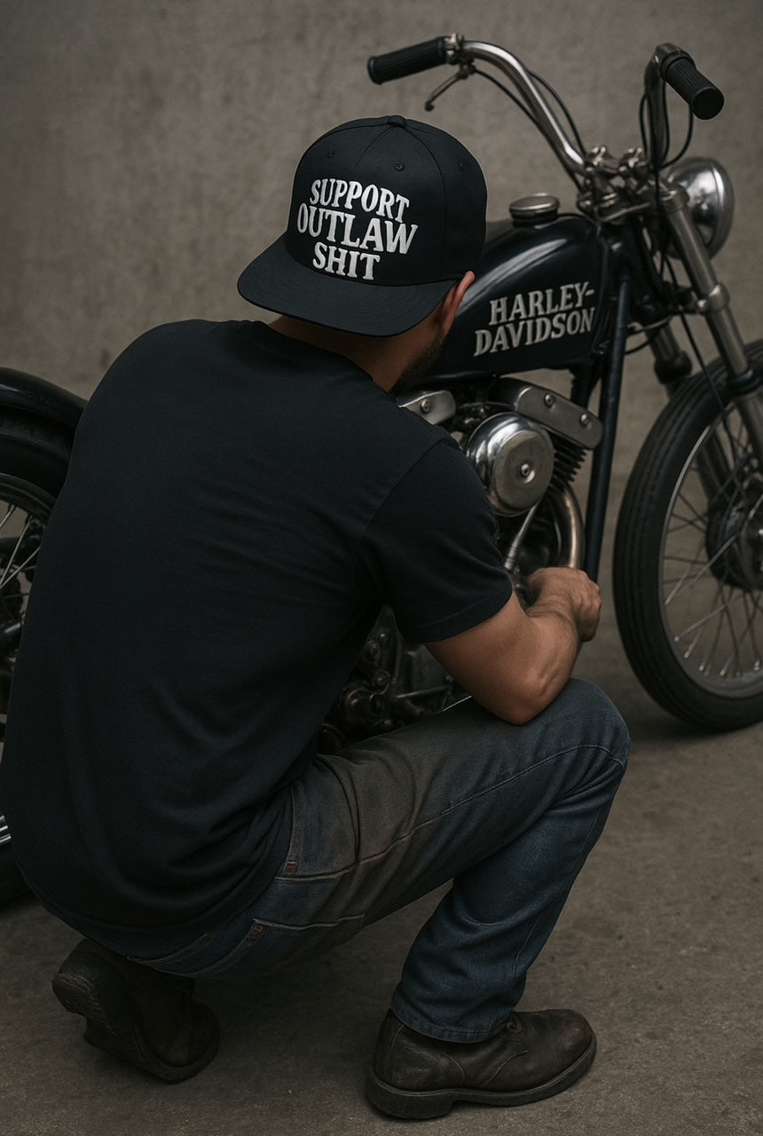 A black snapback hat with a curved brim, featuring a bold embroidered patch that reads "Support Outlaw Shit" in white lettering. The hat has a structured, six-panel design and is styled with a rugged, rebellious aesthetic. Branded by Highway Division, it's shown against a neutral background with subtle lighting to highlight its texture and detail.