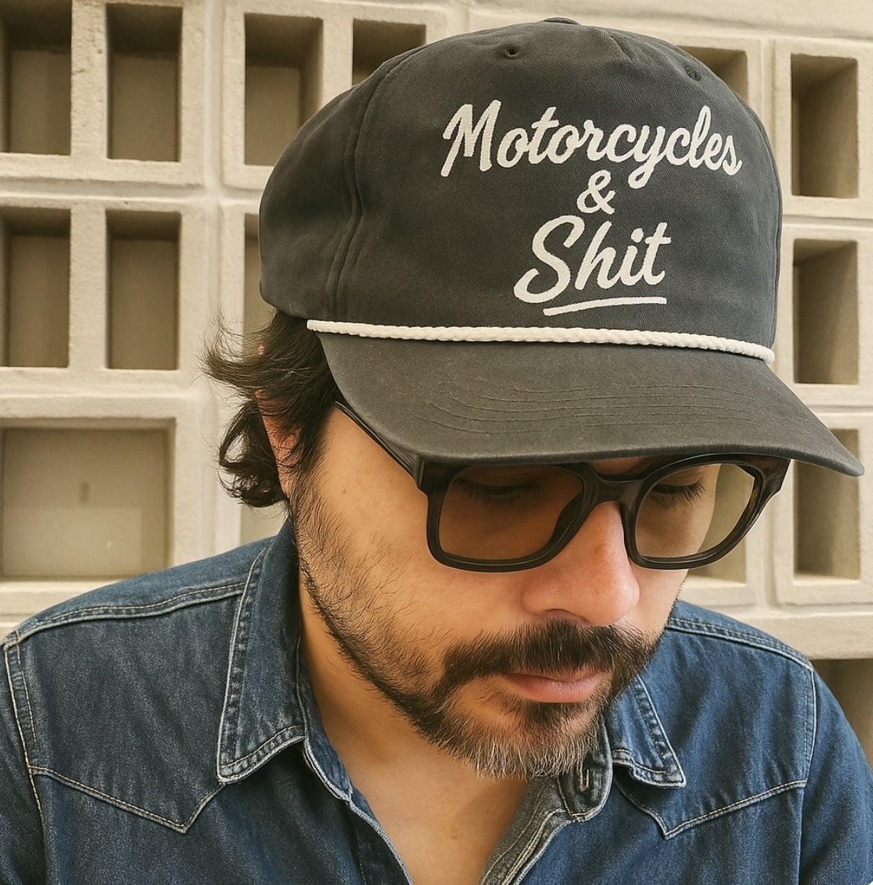 Black 'Motorcycles and Shit' trucker hat by Highway Division, styled with biker streetwear, worn by a rider standing next to a custom Harley-Davidson motorcycle.