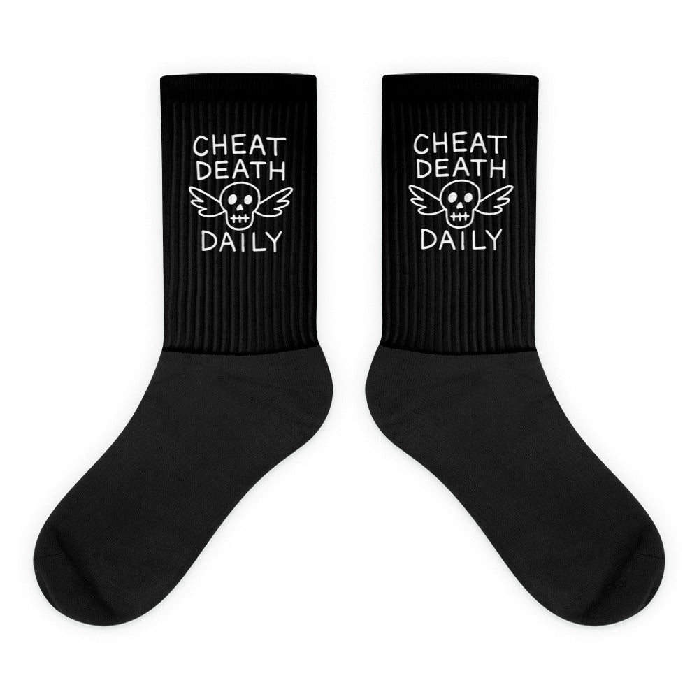 A pair of black crew-length socks from Highway Division is displayed either worn by a model or laid flat on a neutral surface. The socks feature bold, white woven text that reads “CHEAT DEATH DAILY” across the upper cuff area, positioned to be visible above most shoes or boots. The message is aggressive, fearless, and consistent with the brand’s rebellious, no-compromise aesthetic. Made from a soft yet durable cotton blend, the socks have a ribbed texture that ensures a snug, stay-up fit. The heel and toe a