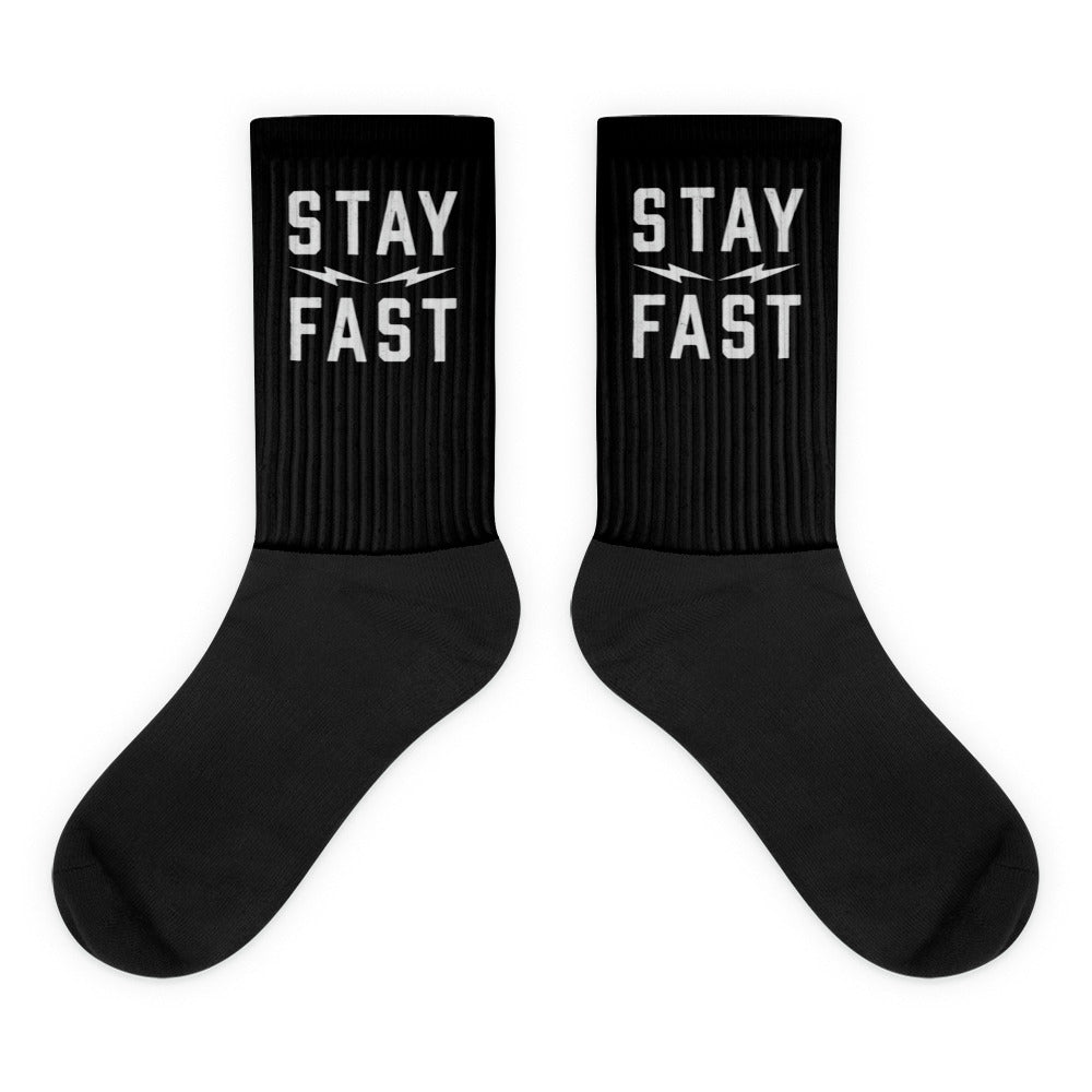 A pair of black crew-length socks from Highway Division is displayed on a clean, neutral background or worn by a model styled in rugged, streetwear-inspired clothing. The socks feature bold white woven text across the top cuff that reads “STAY FAST” in all capital letters. The lettering is clear, sharp, and intentionally placed to remain visible just above the ankle when worn with sneakers or boots, making a bold statement with every step. The socks are made from a high-quality cotton blend 